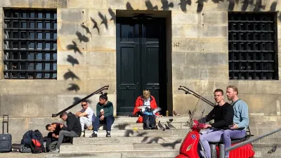 At the backside of the palace, a lot is going on. There is a big black door, and on each side windows with iron cross bars. A 5 layer stone staircase leads up to the door. On the left, 4 tourists are having lunch, each sitting on a different level. The lowest one bends over, the second one a bit, the third sits more or less straight and notices me filming. The forth is a woman. She looks at the other three guys. On the right side is a red scooter, with two young guys on them. 
In the middle is an old man. He wears a crown. He is feeding the doves that sit on his lap.
There is a dark trail of the shadow that dove threw onto the palace, while landing to be fed by the old man.