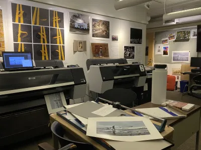 Several big printers in a windowless room. No less than 4 copies of the photo of the woman between the seagulls are scattered around the room.