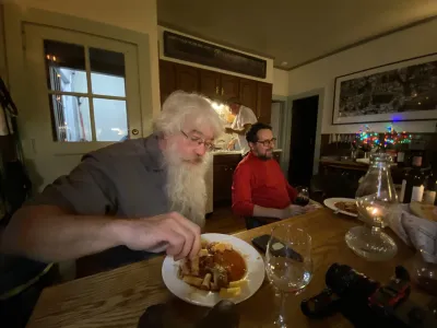 Wide angle shot of a man with a long white beard and longish hair. It's Pete Mauney. He has a very deep voice. He reaches for his food, exaggerated with by the wide angle lens. In the back, in red sits another man, with black hair.  That's Matt Malsky.