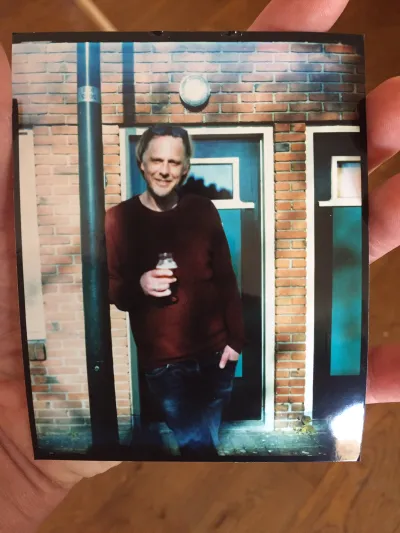 My hand holding a 4x5 inch photo by the edges, of a guy in his forties in front of his house, leaning into a lamp post, holding a beer. The colors seem off and correct at the same time.