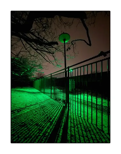 I said the top photo was the best, but I work the hardest on this one. Again a standing photo. It shows the fence coming into the frame on the right, exiting into the dark distance near the left edge. The green lamp is hidden behind poles of the fence. On green lit snowy floor, we see a diverging pattern of black shades. The top half of the image is the dusty rose sky, cut by thick black branches from the top left, that are brighter green the smaller they get. More or less in the middle is a lamp post. Its lamp is like a flying saucer, between the branches. It’s out of order, but its glass is lit by the green flood light. This makes the glass into a green object in the dusty rose sky. It may sound like simple photo, but the lines in the green bottom half and the naturel items + green UFO in the top half against the dusty rose sky make it quite strange.