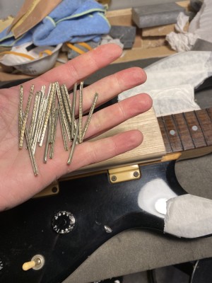 A bass lies on a workbench. The frets are off, the gaps they left are filled with fine strips of wood. A hand comes into the frame, showing the 22 frets. They have been bent. 
