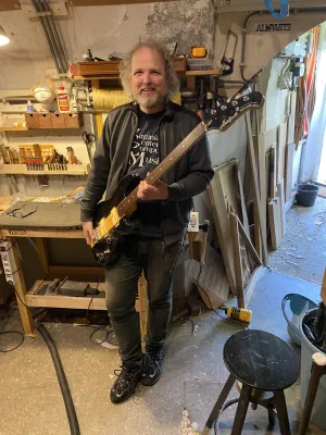David standing in the workshop. Tools behind him. The workshop is in a Utrecht Werfkelder, and you can see the ceiling is curved like a tunnel. Daylight is coming in from the right. Happy David is happy with the finished bass.