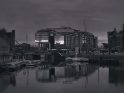 A dark image. In the center is a huge modern hotel building that dominates the image. There is a textured overcast sky. To the left is a bit of the late 19th century central station, to the right some typical 17th century Amsterdam canal houses. The front of the image is water. There are 5 tourist boats in it, the size of city busses, but low enough to crawl underneath the bridges. Everything is dark grays. But in the modern hotel, the heat isolation of the HR glass does reflect IR very well. We can see a bright sunset mirrored in it. The reflection of the building in the water does not show this sunset, likely because of the viewing angles. The result is slightly distopic.