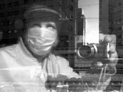 Selfy in a train dirty window with a complex layered background. You can see the camera in my hand. I'm wearing a surgical mask and a hat.