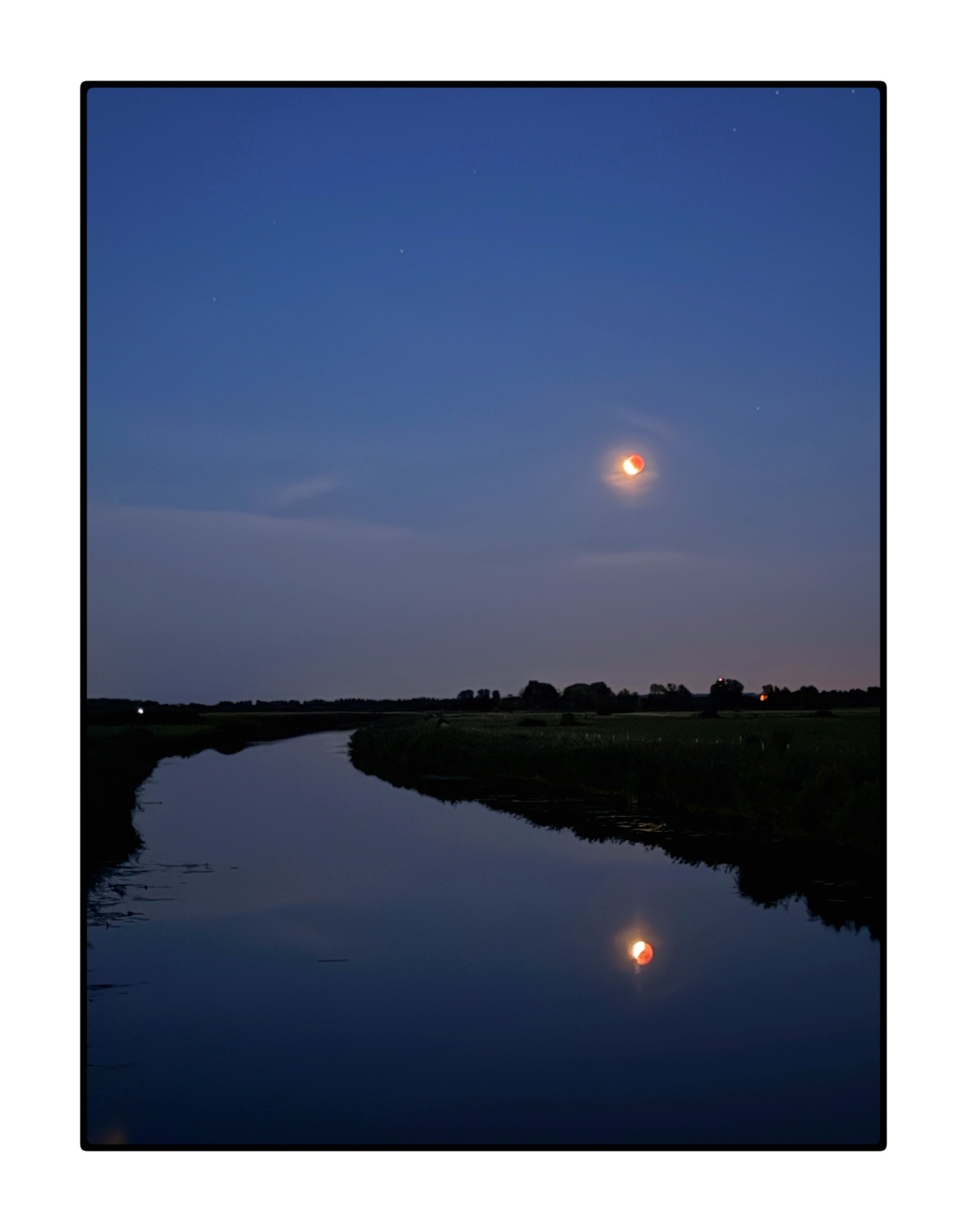 Het is het begin van de nacht. De lucht is diep blauw. Een brede sloot leidt naar de horizon, links en rechts zijn de kades heel donker. Een paar lichtjes aan opvallend vlakke de horizon. De lucht weerkaast in de sloot. Het water is vlak. De maan is vol, maar 2/3e verduisterd. Hij reflecteert ook in de sloot. De staande foto komt grafisch over.