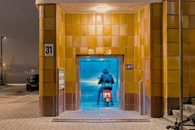 Een brommerrijder parkeert in de lift die naar de Maas-tunnel leidt. Naast het liftgebouw zien we nog net haven in de mist, en een stukje van een auto die om de hoek lijkt te gluren. De hele foto is gelig, door natrium SOX lampen en gele tegels op de muren. Maar in de lift is het licht blauw, om te voorkomen dat junkies er gaan injecteren.