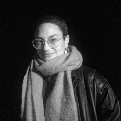 Portret van Yara op 6x6 film. Kort haar, grote rode bril, wollen sjaal om en leren jas. Ze zit iets links in het frame. De achtergrond is zwart. 