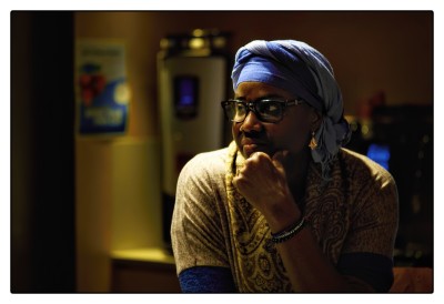 Een vrouw met een blauwe doek op haar hoofd gele kleding is theatraal belicht, maar zit, aan de koffiemachine achter haar te zien, in een soort kantine. Ze heeft haar hand op haar kin en kijkt observerend naar links. Ze is niet helemaal aanwezig. Het herinnert aan het meisje met de parel van Vermeer.