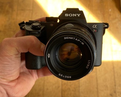 My hand holding a Sony camera, with a Sovjet lens on it. Zenitar is spelled in Cyrillic. The lens it tilted a bit to the left. 