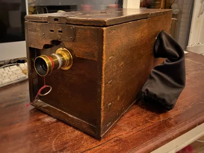 The box camera, on a wooden table, both dark brown stained wood. On the front, a lens in a brass barrel. On the side, a 30 cm sleeve black sleeve comes out. The end has elastic bands in it, like a changing bag. The box still looks as antique as it is.