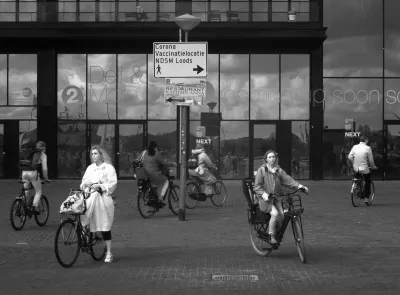 4 people on bikes arriving at NDSM, two others waiting to get on that ferry to go to Amsterdam Centraal. Especially a lady on the left with a bright long coat and dito shoes, stands out against the gray scales. A bloke on the right with a bright coat balances the image. Clouds are mirrored against a modern building with lot's of coated glass that should keep heat out and in, but let light through. That coating seems to work quite well. There is improvised signage where to get your corona vaccination.