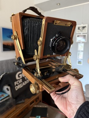 A hand holding up a beautiful wooden camera with leather bellows and brass hardware, the back and front of which are in tilted, swung and shifted position, showing off the upshot of a bellows camera. In the background, a record player. A sweater that says "Go for ya dreams" is draped over a little TV screen.